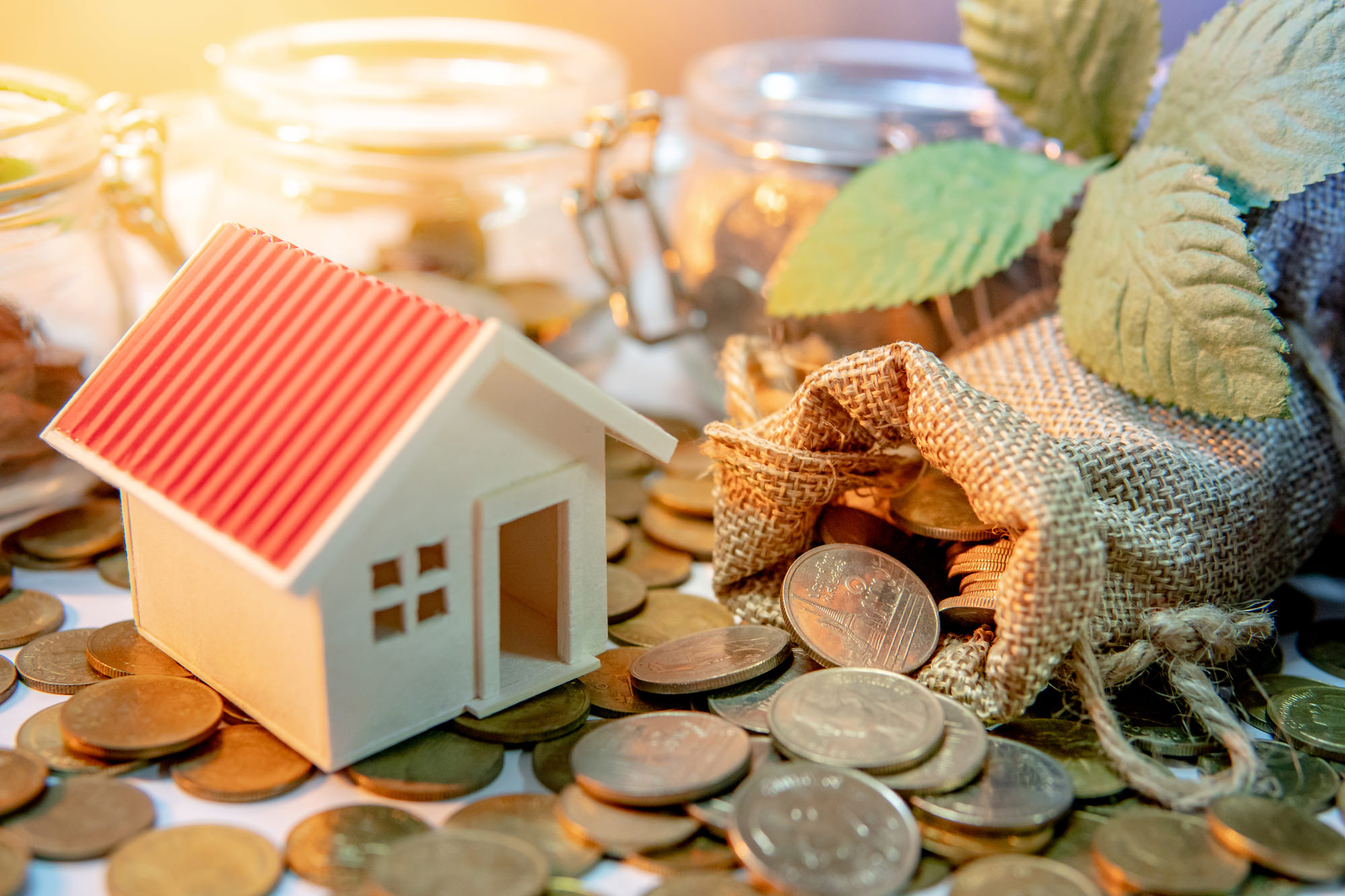 家のミニチュアと貯金箱、硬貨が並ぶテーブル。住宅購入に必要な資金計画を考える場面